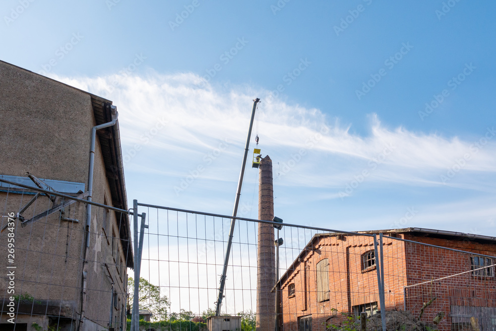 2 men in a basket hanging from a gigantic crane, painstakingly removing a factory chimney