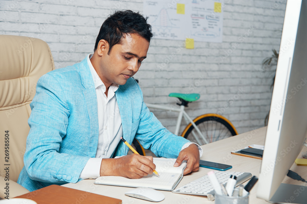 Indian serious professional in blue jacket sitting at office desk and writing future plans in his notepad while working at modern office Indian serious professional in blue jacket sitting at office desk and writing future plans in his notepad while working at modern office