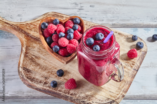 Fresh smoothies in a glass jar with ripe raspberries and blueberries in a wooden heart-shaped bowl on rustic cutting board. Healthy food concept. Dessert for valentines day. Top view