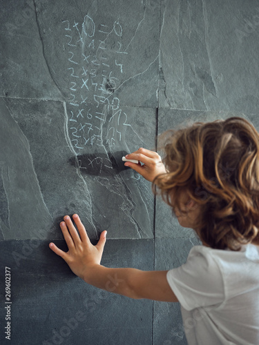 Back view of long hair boy writing by chalk multiplication table on grey stone wall photo