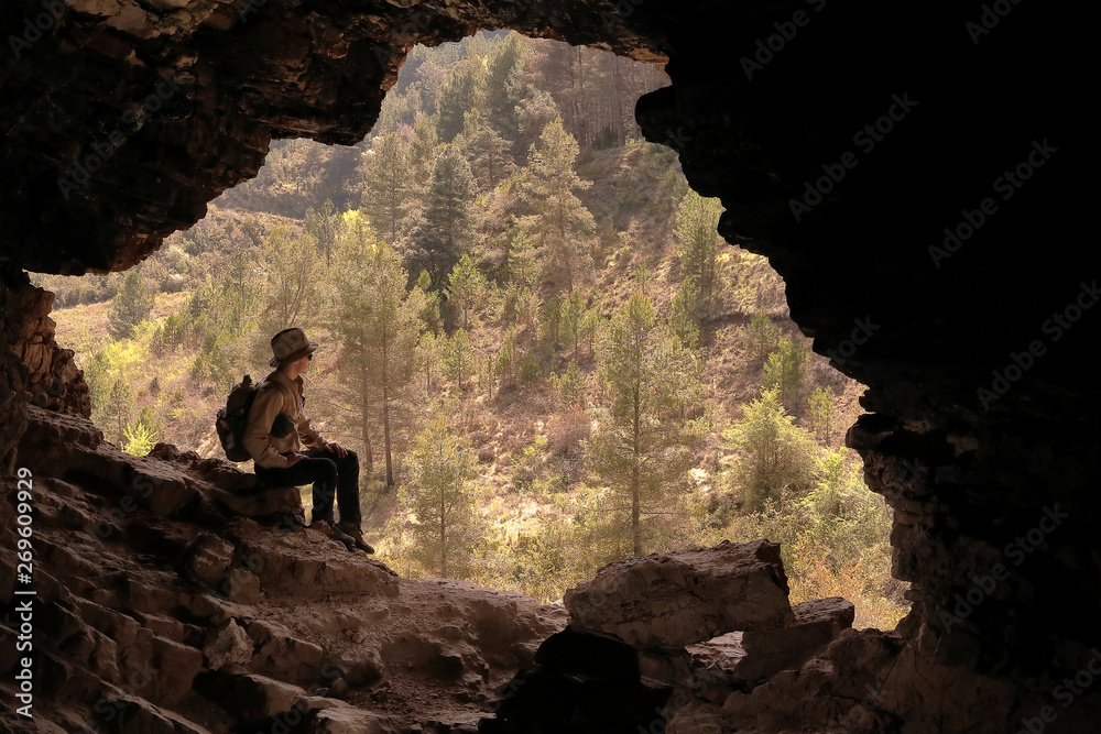 BOY WITH HAT AND BACKPACK SITTING ON A ROCK AT THE ENTRANCE OF A MYSTERIOUS CAVE IN FRONT OF A VALLEY WITH MANY GREEN TREES