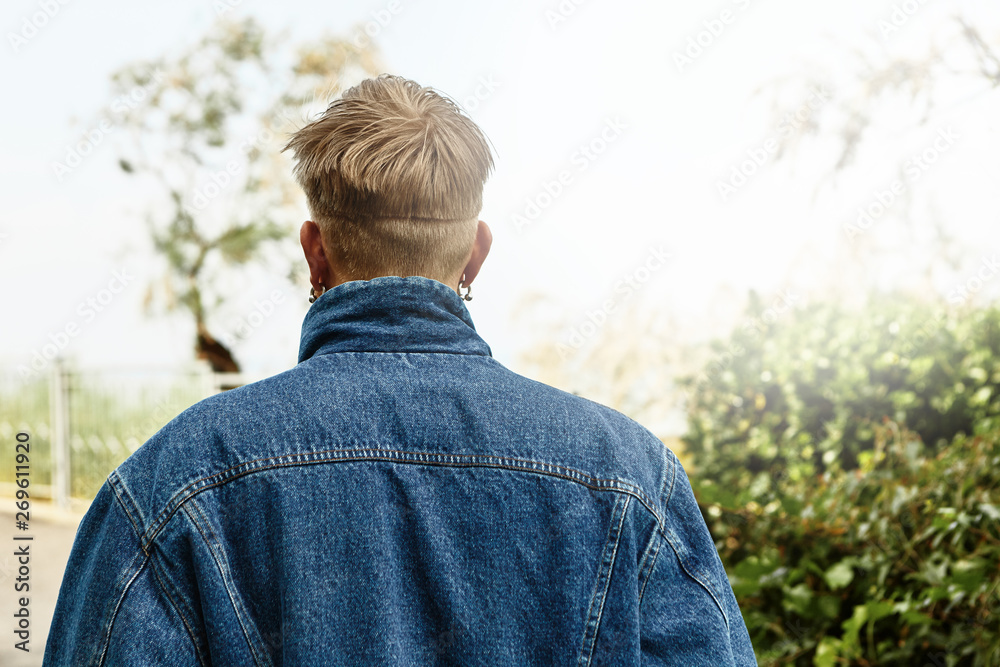 Style, fashion and trends concept. Outdoor rear shot of unrecognizable young blonde man with stylish haircut walking among green trees on sunny day, wearing earrings and stylish jeans jacket