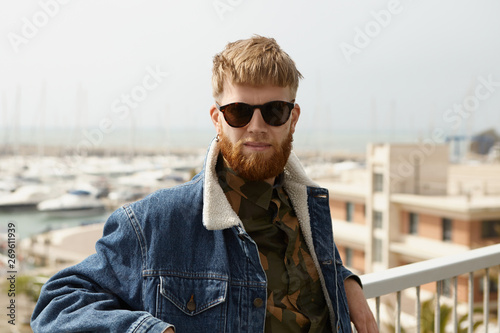 Handsome confident man with fuzzy beard standing on view point with arms on white fence rails, contemplating amazing sea scenery, wearing stylish shades and jeans jacket, enjoying nice summer day