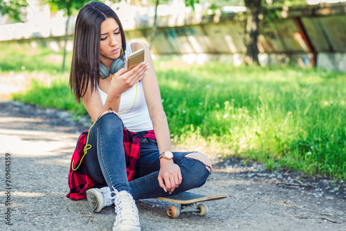 Girl sitting on skateboard and use mobile phone. Outdoors, urban lifestyle..