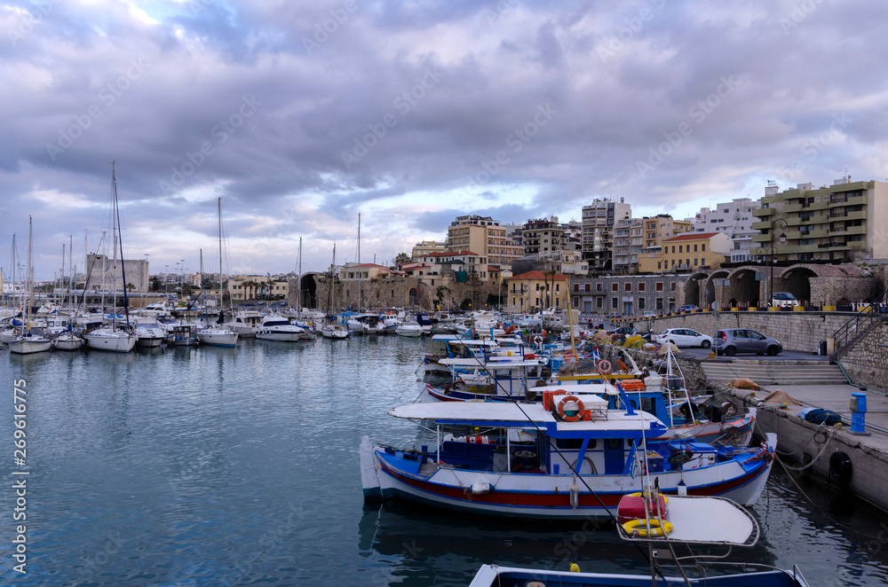 Heraklion, Crete Island - Greece. View to the old Venetian port with the traditional fishing boats and the Heraklion city with the old Venetian shipyards. Sunset, cloudy sky