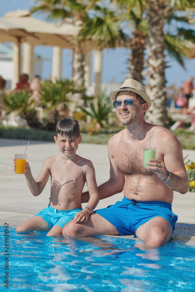 Fototapeta premium The portrait of European man and his son at the hotel’s swimming pool. They are drinking cocktails and enjoying their holidays.