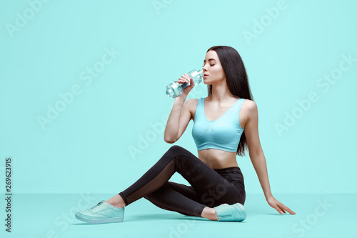 Amazing woman in trendy sportswear drinking water from bottle to stay hydrated after workout. Beautiful slim brunette young girl in fashion leggings and top on blue background.