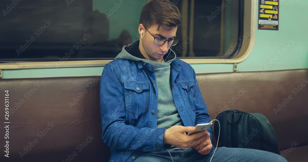 Fashionable brutal guy in glasses and headphones rides in an empty subway car. Young man sits in the subway and presses in the phone.