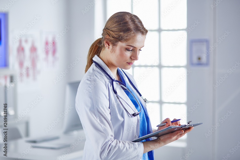 Young woman doctor is standing with board with clipboard smiling in hospital office Young woman doctor is standing with board with clipboard smiling in hospital office