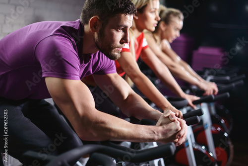 Close up hands of man biking in spinning class. Group of smiling friends at gym exercising on stationary bike. Happy cheerful athletes training on exercise bike.