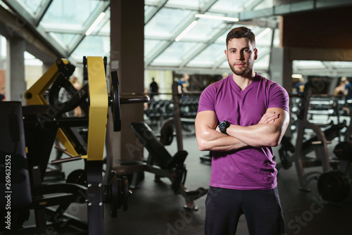 Handsome friendly personal trainer in gym. Young sporty man smiling standing on fitness club background