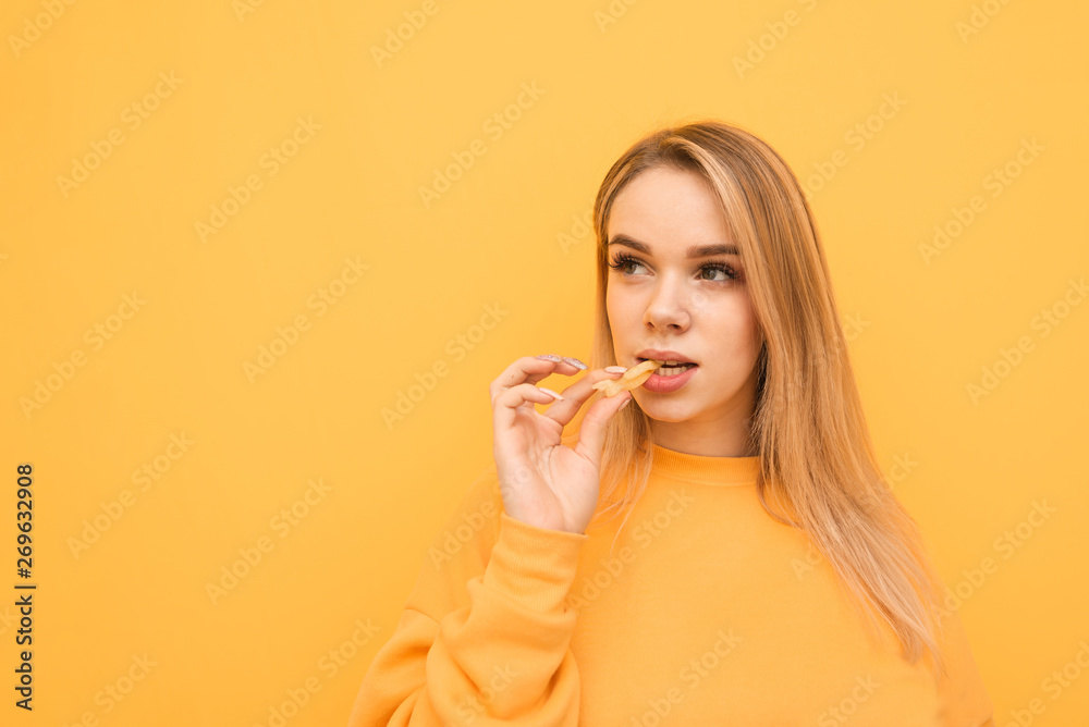 Portrait of an attractive girl with french fries in her mouth with a serious face looking sideways on a lively background. Sweet blonde eating French fries, isolated. Copy-space Portrait of an attractive girl with french fries in her mouth with a serious face looking sideways on a lively background. Sweet blonde eating French fries, isolated. Copy-space