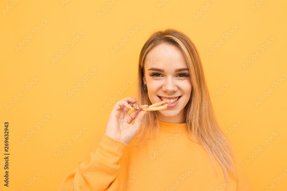 Close-up portrait of a smiling blonde on a yellow background with French fries in his hand, eats and looks into a camera with a positive face. Girl eats fried potatoes and laughs. Close-up portrait of a smiling blonde on a yellow background with French fries in his hand, eats and looks into a camera with a positive face. Girl eats fried potatoes and laughs.
