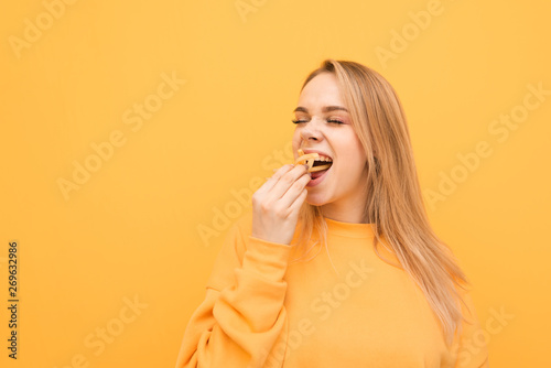 Hungry attractive girl eats a lot of French fries, holds a handful of snacks and puts it in the mouth. Close-up portrait of a girl greedily eats French fries on a yellow background