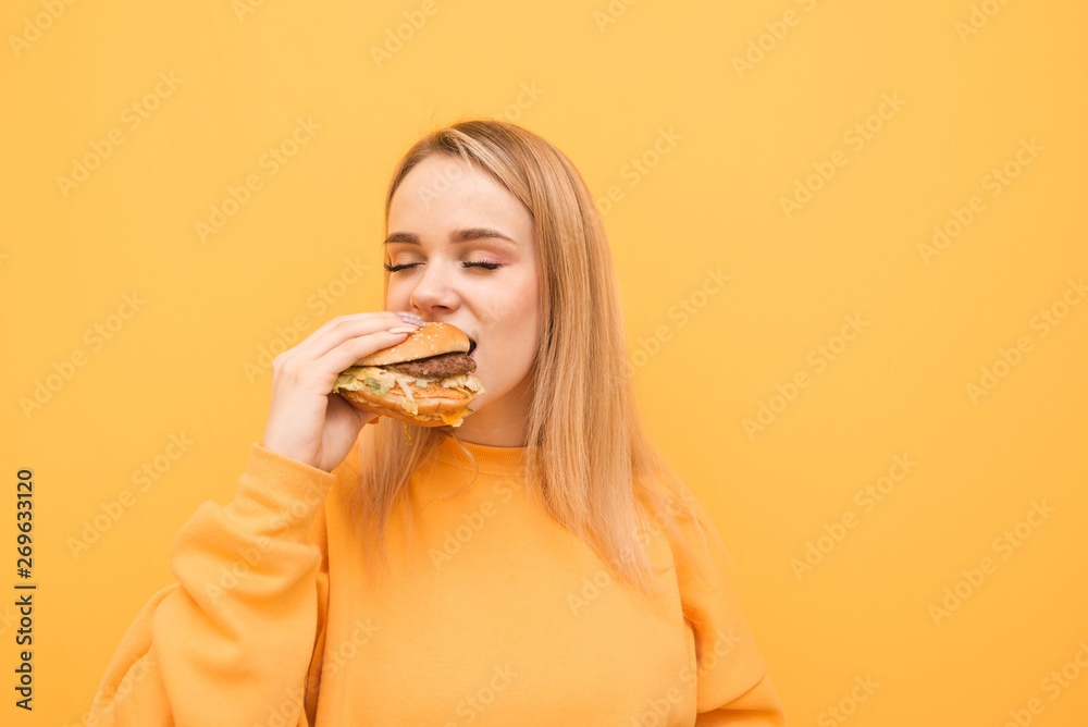 Cute blonde bites a burger with her eyes closed on a yellow background, wearing an orange casual wear, likes to eat fast food.Lady eats bad food,with a pleasing bite of appetizing delicious burger Cute blonde bites a burger with her eyes closed on a yellow background, wearing an orange casual wear, likes to eat fast food.Lady eats bad food,with a pleasing bite of appetizing delicious burger