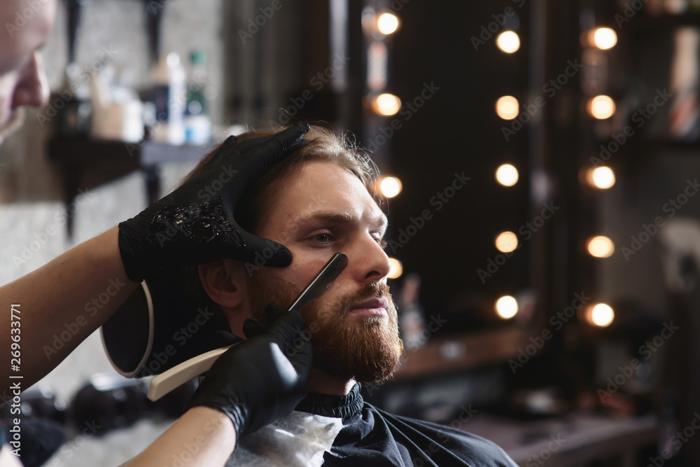 Closeup of barber shaving man beard with straight steel razor blade at barbershop. Brutal hairdresser at work
