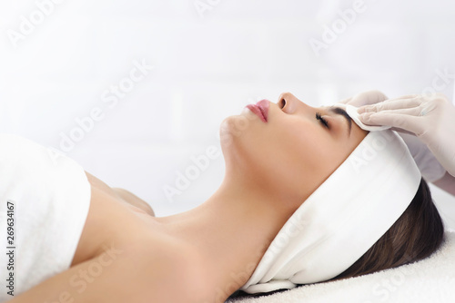 Close up of hands of skillful beautician cleaning and touching female face with cotton pad or sponge. The woman is lying and relaxing. Her eyes are closed with pleasure