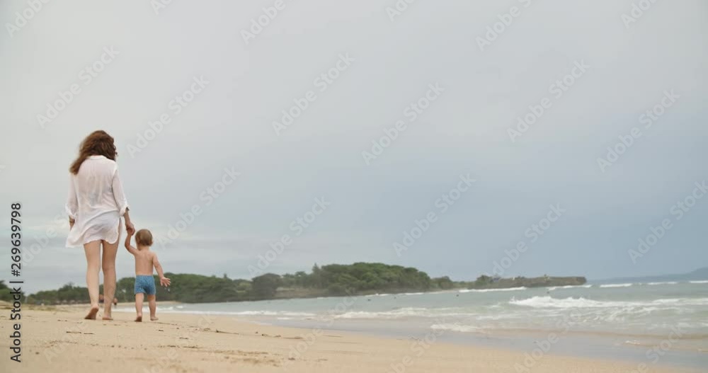 Mother teaching her little baby to walk on sandy beach. Little kid making careful steps, waves of ocean touching their feet - parenthood and childhood concept slow motion 4k Mother teaching her little baby to walk on sandy beach. Little kid making careful steps, waves of ocean touching their feet - parenthood and childhood concept slow motion 4k