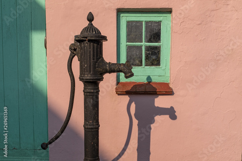 An old fashioned water pump against a pink wall with a green window, with a strong shadow of the pump, nobody in the image