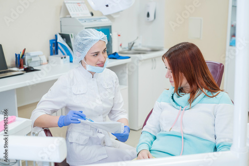 Young beautiful female dentist talking to a patient in the office. A red-haired woman sits in a chair with an orthodontist and listens to appointments. Dental services