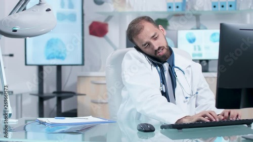 Doctor in his modern office talking on the phone while typing on the computer. CT or MRI 3D brain scans are displayed in the background on big TV and dual monitors