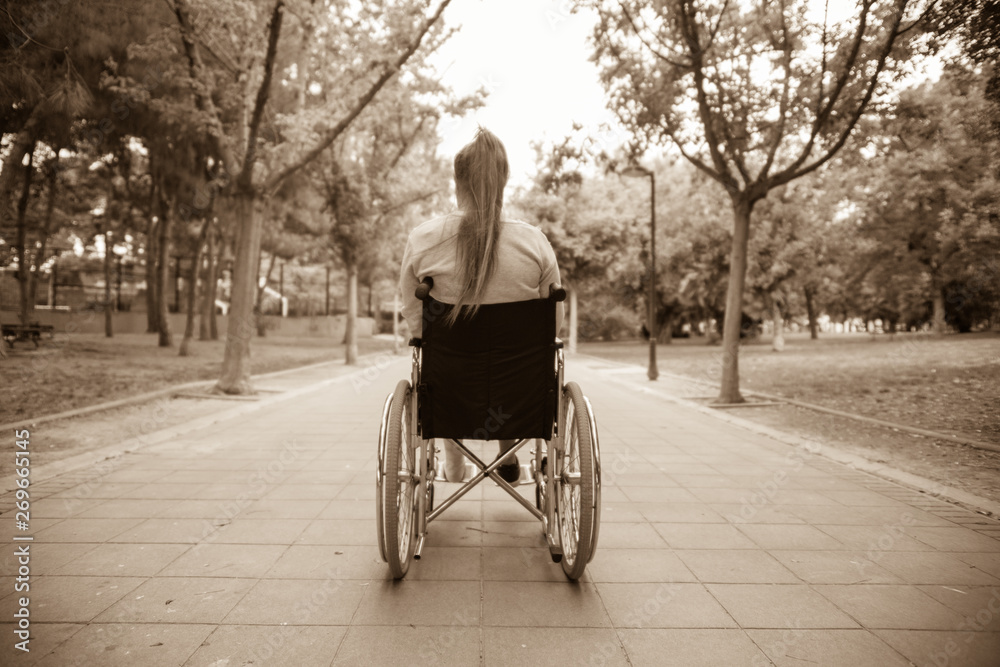 Young girl in wheelchair seen from behind. Outdoors, in a park