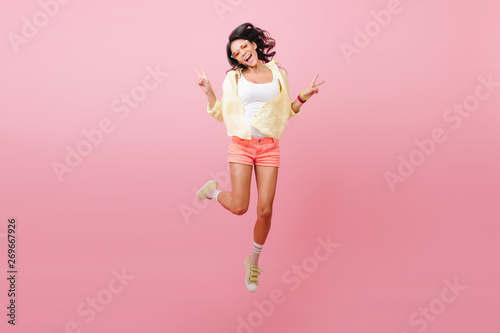 Blissful slim latin girl in casual bright attire enjoying leisure time in studio. Graceful asian woman in sunglasses funny jumping near pink wall.