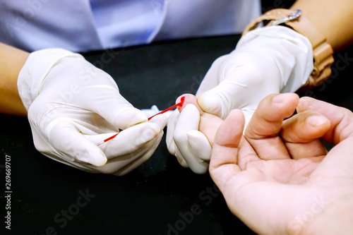 Closeup hands of nurse wear a rubber white medical gloves using capillary tube touch and drain on the blood of human ring finger to check glucose in blood at medical clinic.