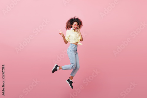 Good-looking lady expressing positive emotions during photoshoot. Glad trendy girl in jeans and sneakers jumping with sincere laugh.
