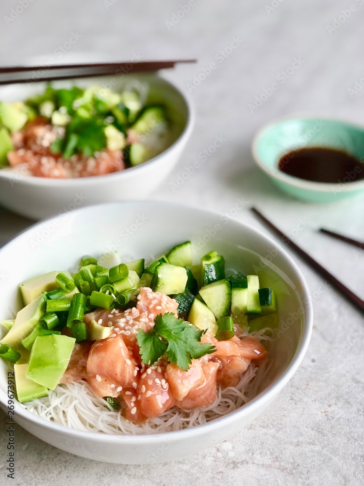 Side view of two Poke bowls with raw marinated salmon, cilantro, avocado, cucumber , sesame seeds and rice noodles served with soy/tamari sauce and wooden chopsticks. Flat lay on light background. Side view of two Poke bowls with raw marinated salmon, cilantro, avocado, cucumber , sesame seeds and rice noodles served with soy/tamari sauce and wooden chopsticks. Flat lay on light background.