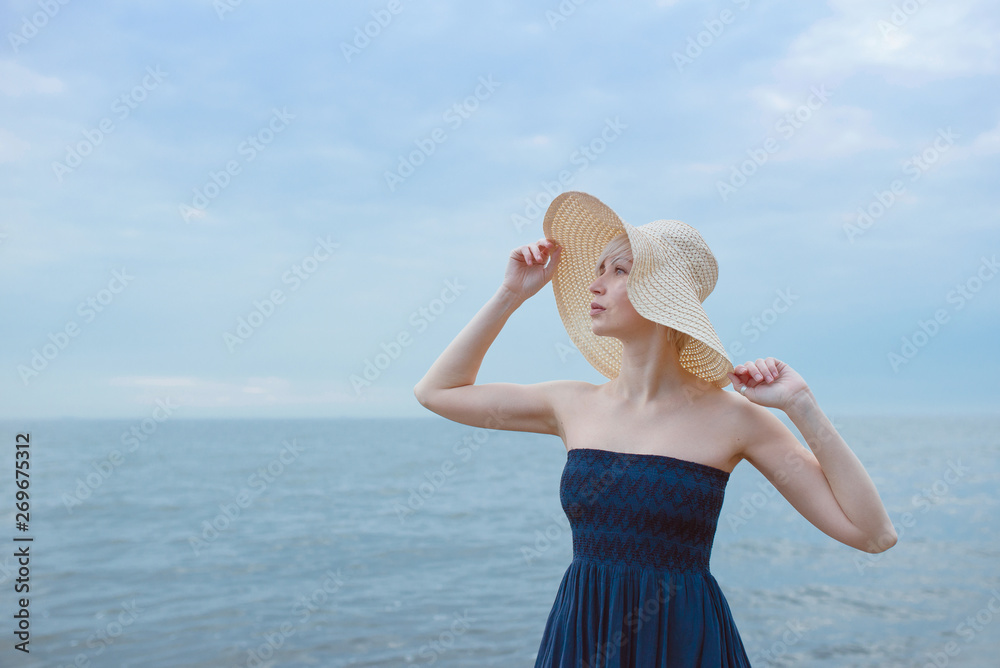 Caucasian beautiful stylish blonde woman in blue dress and in straw hat standing near the sea Caucasian beautiful stylish blonde woman in blue dress and in straw hat standing near the sea