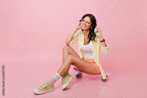 Refined hispanic girl wears yellow sneakers and jacket holding big headphones and laughing. Indoor portrait of cute latin woman with black hair sitting on pink skateboard.