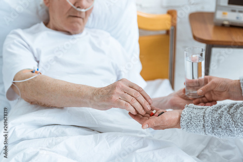 cropped view of senior woman giving glass of water and pills to sick husband in hospital