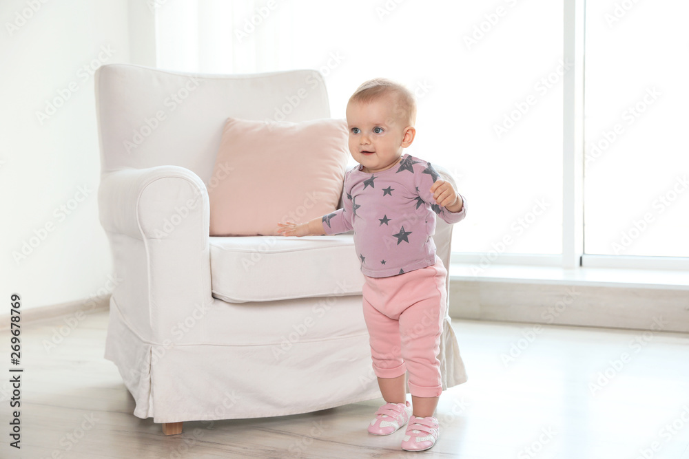 Cute baby girl standing near armchair in room Cute baby girl standing near armchair in room
