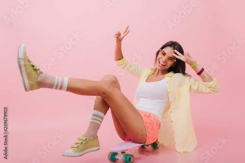 Enthusiastic hispanic girl in trendy yellow jacket sitting on longboard with legs up. Joyful latin woman in colorful bracelets laughing during photoshoot in pink studio.
