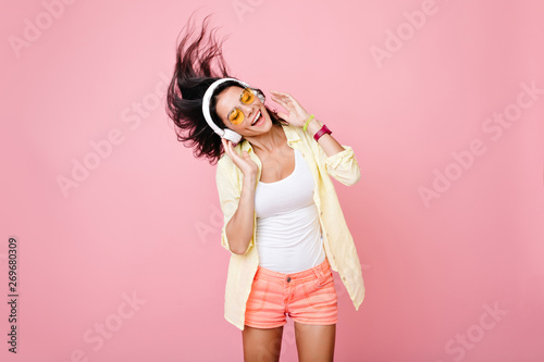 Gorgeous brunette lady in yellow glasses listening music in headphones and singing on pink background. Charming girl in shorts and earphones dancing with hair waving and eyes closed.