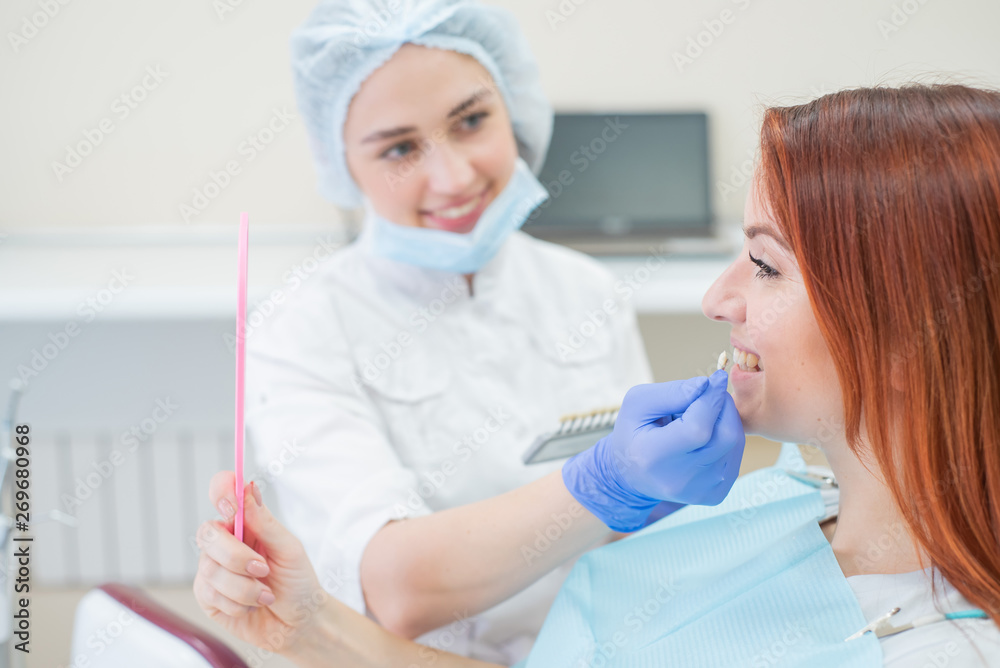 Check and select the color of the teeth in the dentists chair. Dentist makes the process of treating a beautiful young redhaired girl. Selection of a dental implant. Patient looking in the mirror.