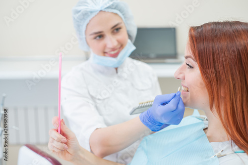 Check and select the color of the teeth in the dentists chair. Dentist makes the process of treating a beautiful young redhaired girl. Selection of a dental implant. Patient looking in the mirror.
