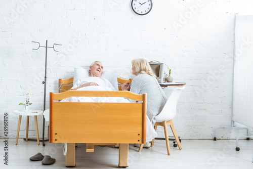 worried senior woman sitting near ill husbend and holding his hand in hospital photo