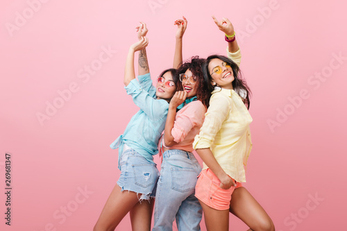 Glamorous hispanic woman in yellow shirt enjoying funny dance with friends. Indoor portrait of three cheerful girls chilling during photoshoot.