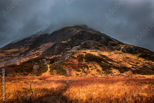 A moody view of a rocky mountain in Glencoe, with red and orange grasses and ferns from the valley floor. Scotland.