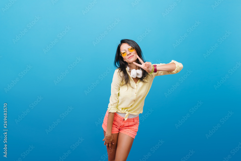 Sporty tanned girl in denim shorts posing on bright blue background with pleasure. Indoor portrait of magnificent european woman in street outfit wears big headphones. Sporty tanned girl in denim shorts posing on bright blue background with pleasure. Indoor portrait of magnificent european woman in street outfit wears big headphones.