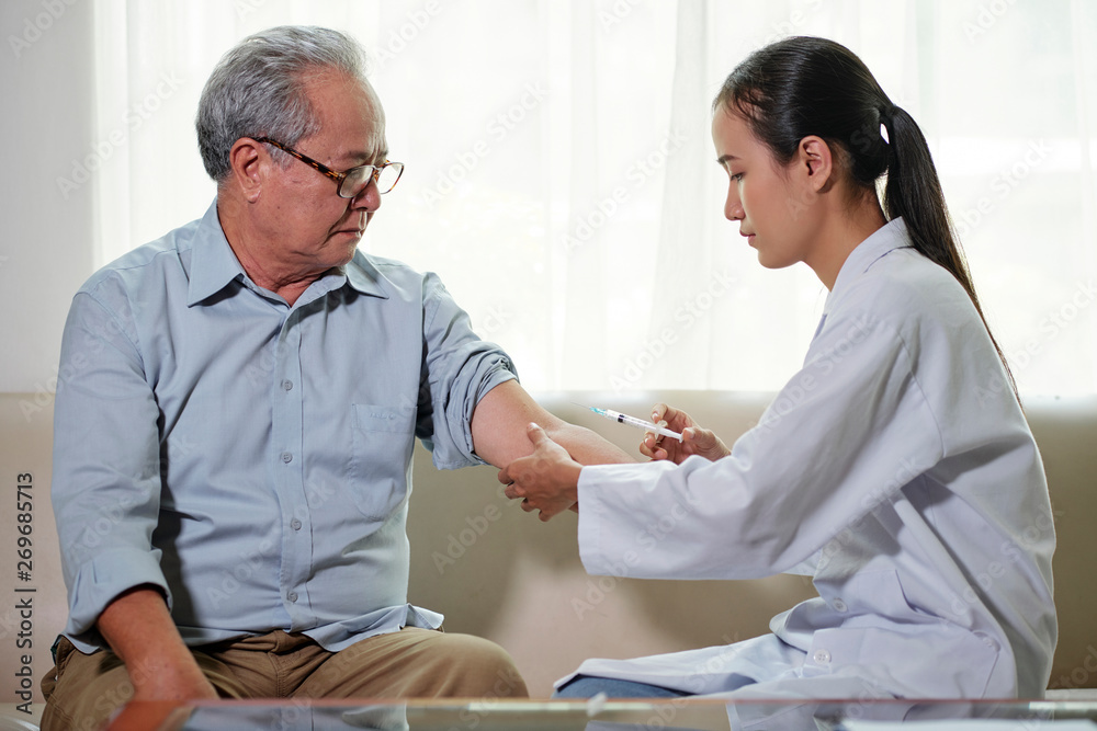 Obraz premium Asian young doctor in white coat sitting on sofa and making an injection to senior man while they sitting in the living room