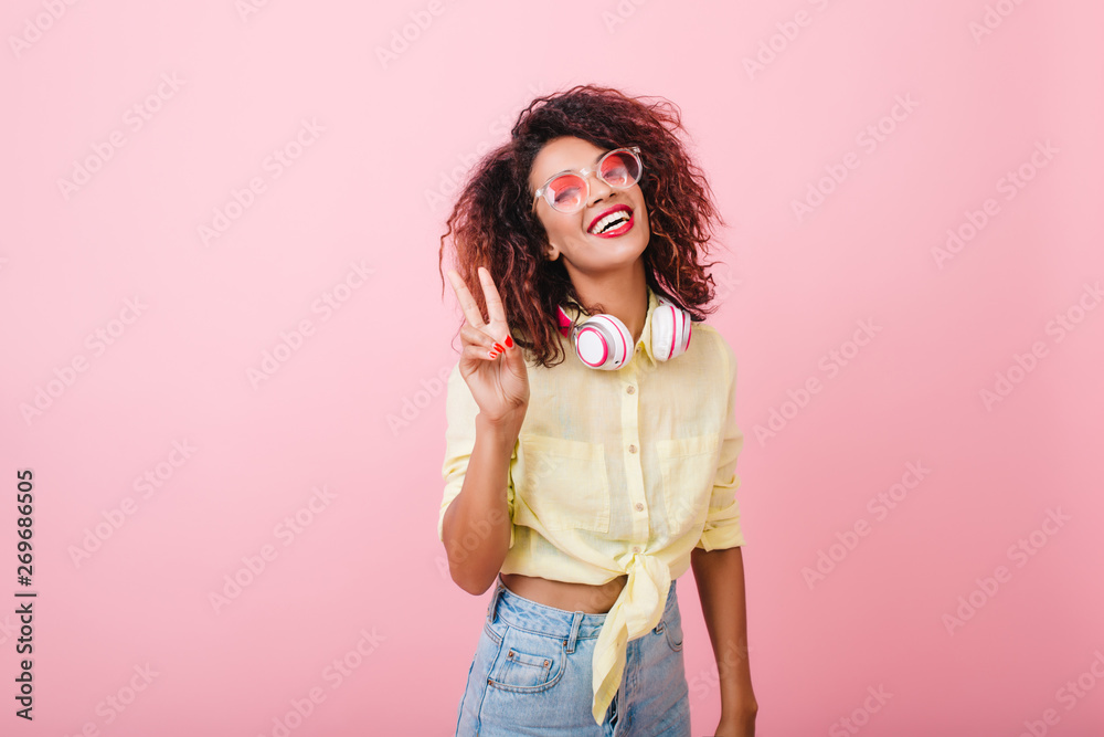 Pleased young female model in summer shirt laughing during indoor photoshoot. Sincere african girl with curly hair relaxing in studio with pink walls. Pleased young female model in summer shirt laughing during indoor photoshoot. Sincere african girl with curly hair relaxing in studio with pink walls.
