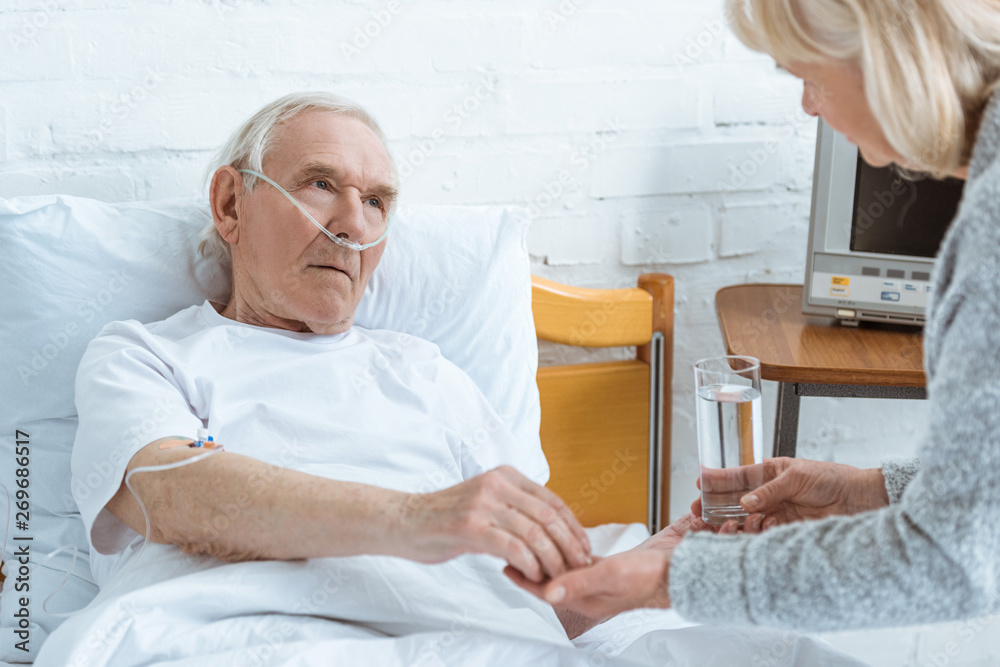 Obraz premium cropped view of senior woman giving glass of water to sick husband in hospital