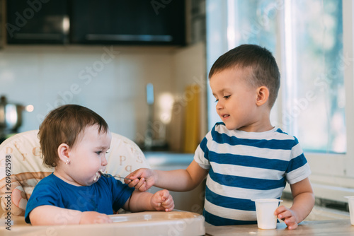 Kids in the kitchen, brother feeds baby sister yogurt with a spoon, she helps parents