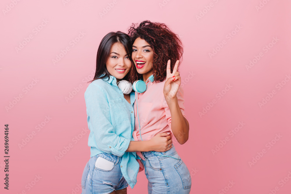 Pretty european woman with short black hair gently embracing african female friend in pink shirt. Slim curly lady huggs brunette caucasian girl during photoshoot. Pretty european woman with short black hair gently embracing african female friend in pink shirt. Slim curly lady huggs brunette caucasian girl during photoshoot.