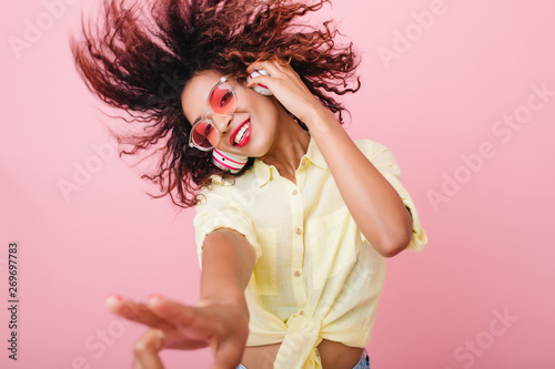 Close-up portrait of adorable curly girl happy smiling during photoshoot. Stunning african woman with light-brown skin relaxing in headphones and funny dancing on colorful background.