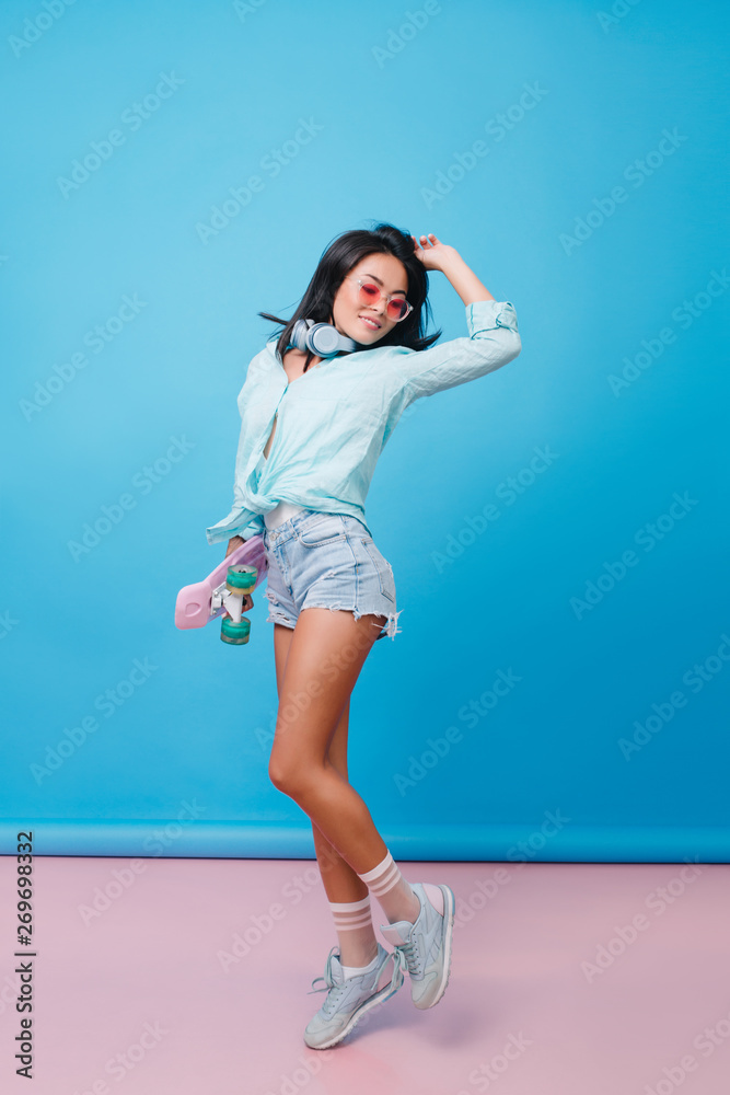 Full-length portrait of slim hispanic woman expressing happy emotions in blue studio. Sensual latin girl with tanned skin in street outfit dancing with hand up.