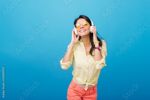 Inspired asian female model in pink wristwatch and green bracelet listening music on blue background. Indoor photo of ecstatic latin girl in orange sunglasses touching headphones and smiling.